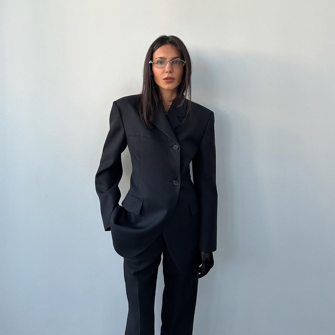 An adult woman with long dark hair and glasses poses in a black pantsuit against a white wall. Minimalist fashion portrait in studio.