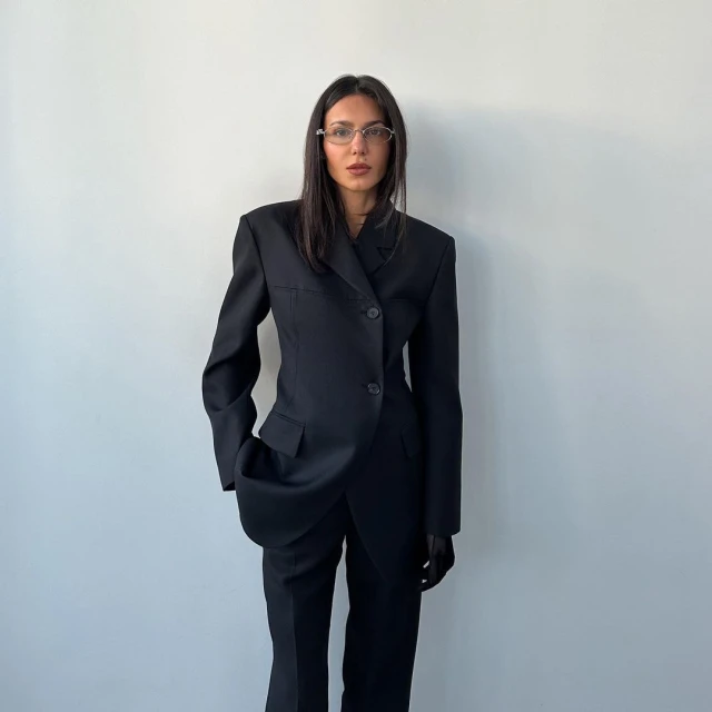 An adult woman with long dark hair and glasses poses in a black pantsuit against a white wall. Minimalist fashion portrait in studio.