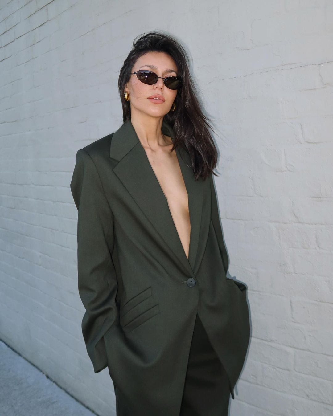 A stylish woman with long dark hair and sunglasses poses in an olive green suit against a pale brick wall, exuding fashion-forward confidence.