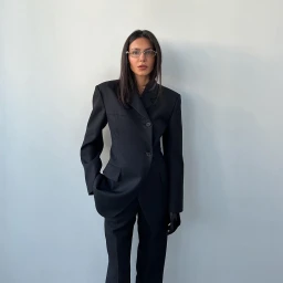 A woman wearing a tailored black blazer and matching trousers stands against a plain white wall.