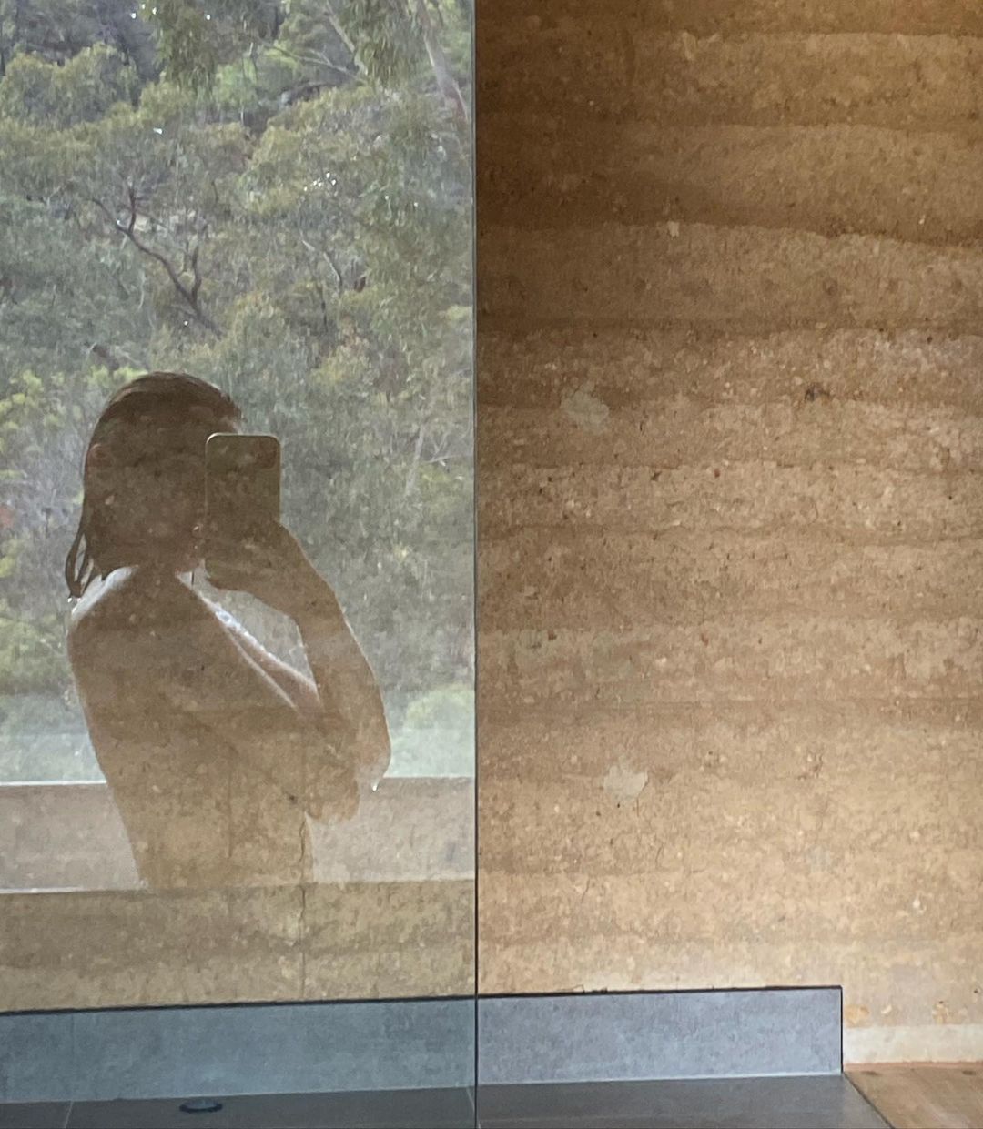 Indoor shot of a person in a bathroom, reflected in a glass shower wall with outdoor trees and a wall with stacked earth.