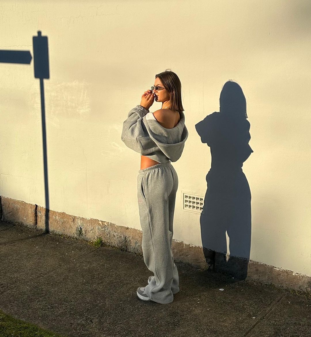 A woman in a grey hooded sweatshirt and sweatpants stands in full sunlight against a white wall, casting strong shadows on the surface.