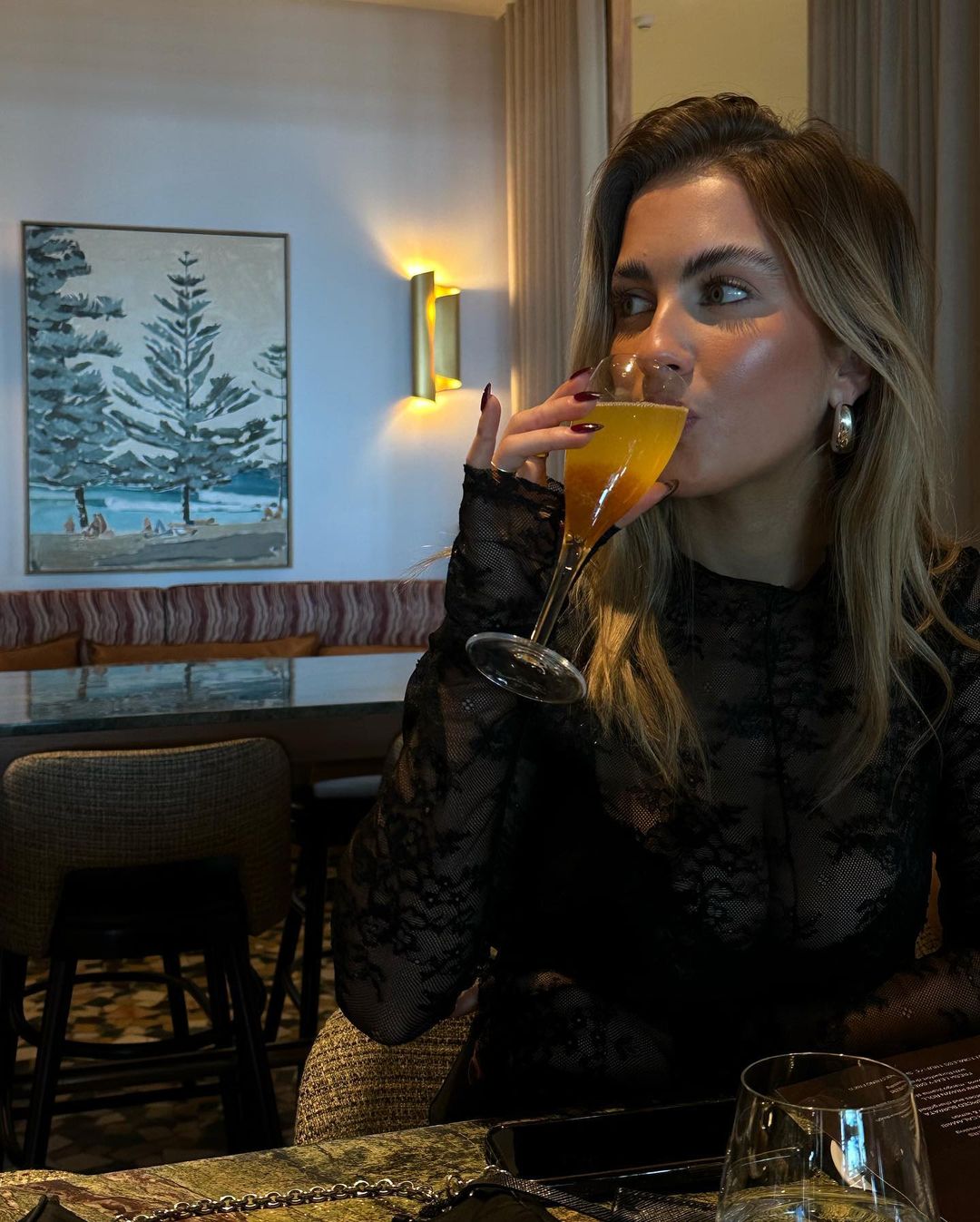Blond woman in black lace dress drinks an orange cocktail in a restaurant with beach painting and warm lighting. Dining experience.
