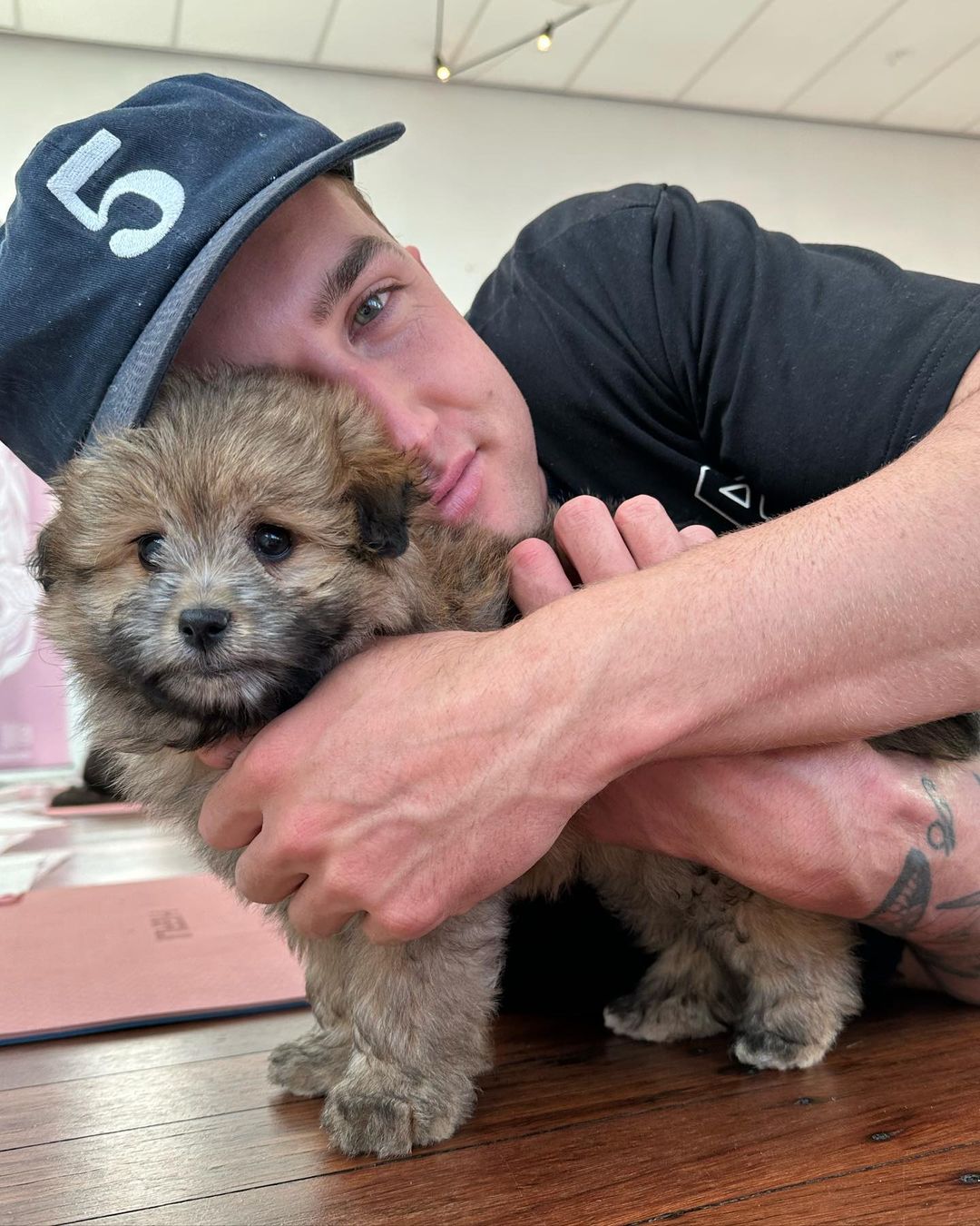 A man in a baseball cap embraces a fluffy brown puppy, set against a softly lit indoor background with wooden floors.