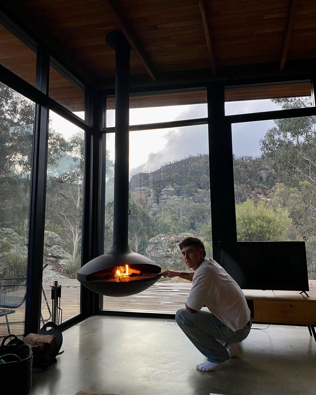 Adult male tending a modern hanging fireplace in a contemporary home with floor-to-ceiling windows showing an outdoor mountain landscape.