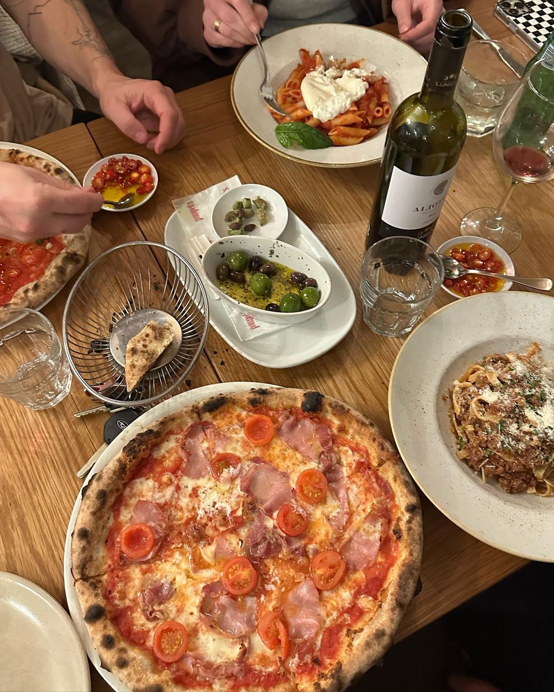 Overhead shot of a wooden table filled with Italian food, including two pizzas, pasta dishes, a wine bottle, and assorted appetizers.