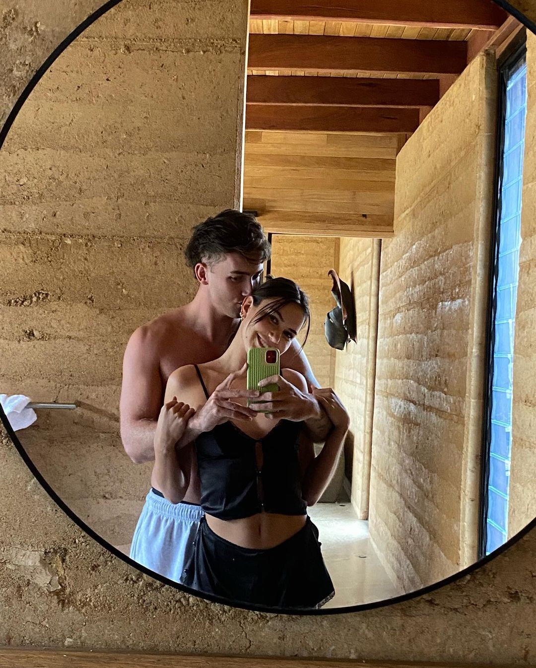 A couple poses for a selfie in a bathroom featuring a textured wall, with a man embracing a woman holding a smartphone.