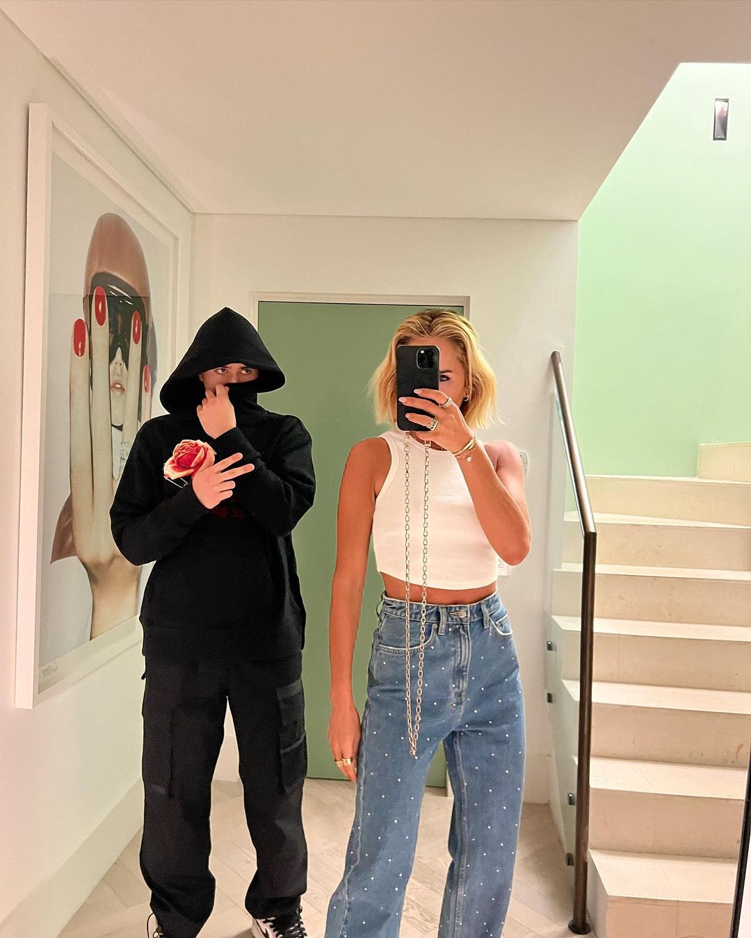 Two people in a hallway, one in a hooded sweatshirt with a rose graphic and the other in a white tank top and jeans, with artwork visible.