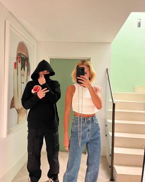Two people in a hallway, one in a hooded sweatshirt with a rose graphic and the other in a white tank top and jeans, with artwork visible.