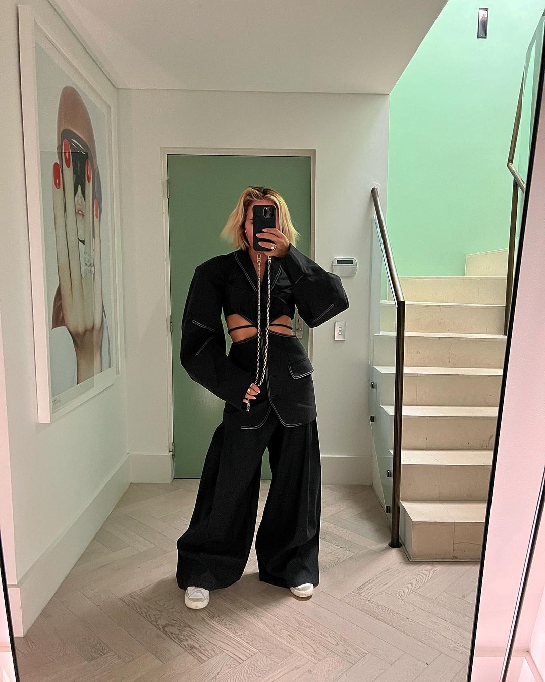 Woman in a black deconstructed suit takes a mirror selfie inside a modern home with white walls, herringbone floors, and a lime-green accent wall.