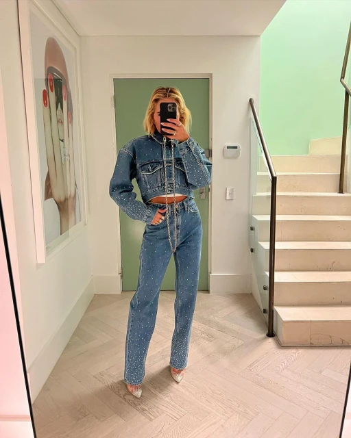 A woman in a denim outfit takes a mirror selfie in a modern home. She's posing near a staircase with an art piece on the wall.