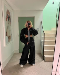Woman taking a mirror selfie in a hallway wearing a black cutout blazer and wide-leg trousers, holding a phone with a chain strap.