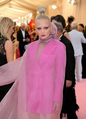 Woman with unique makeup wearing a pink blazer dress and sheer cape poses on a red carpet among other people and flashes from cameras.