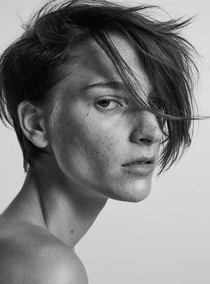 Portrait of an androgynous adult woman with short dark hair and freckles, looking at the camera in a studio setting against a white background.