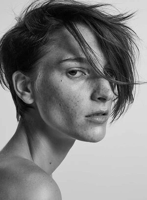 Portrait of an androgynous adult woman with short dark hair and freckles, looking at the camera in a studio setting against a white background.
