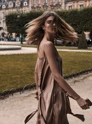 A woman with long brown hair wearing a brown dress poses outdoors in front of green lawns and architecture in an eye-level shot.