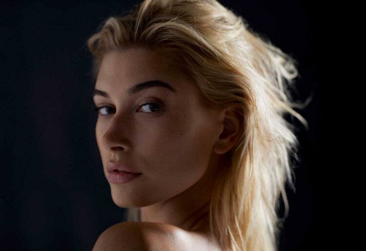 Close-up portrait of a woman with blonde hair looking over her shoulder against a dark background, highlighting her complexion and features.