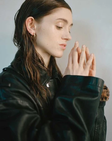 Close-up portrait of a woman with wet dark hair wearing a black leather jacket and small gold hoop earrings against a white background.