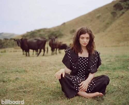 Lorde sits barefoot in a field of grass wearing a polka-dot dress, with cattle grazing and green hills in the distance, under an overcast sky.