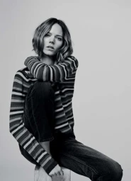 Woman in striped long-sleeve sweater and dark jeans seated against a plain neutral background