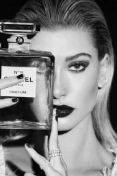 Close-up monochrome portrait of a woman holding a Chanel No. 5 perfume bottle, featuring wet-look slicked-back hair and dark lipstick.
