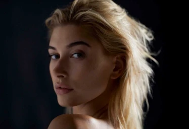 Close-up portrait of a woman with blonde hair looking over her shoulder against a dark background