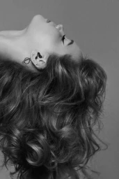 Close-up profile of a woman with voluminous textured curls and winged eyeliner against a grey backdrop