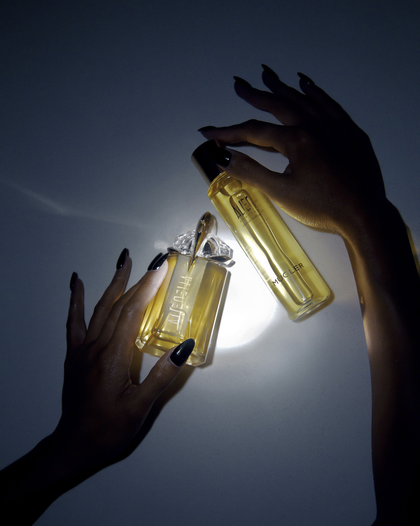 Two glass perfume bottles are held by a person with long black nails against a gradient blue and white background with dramatic lighting.