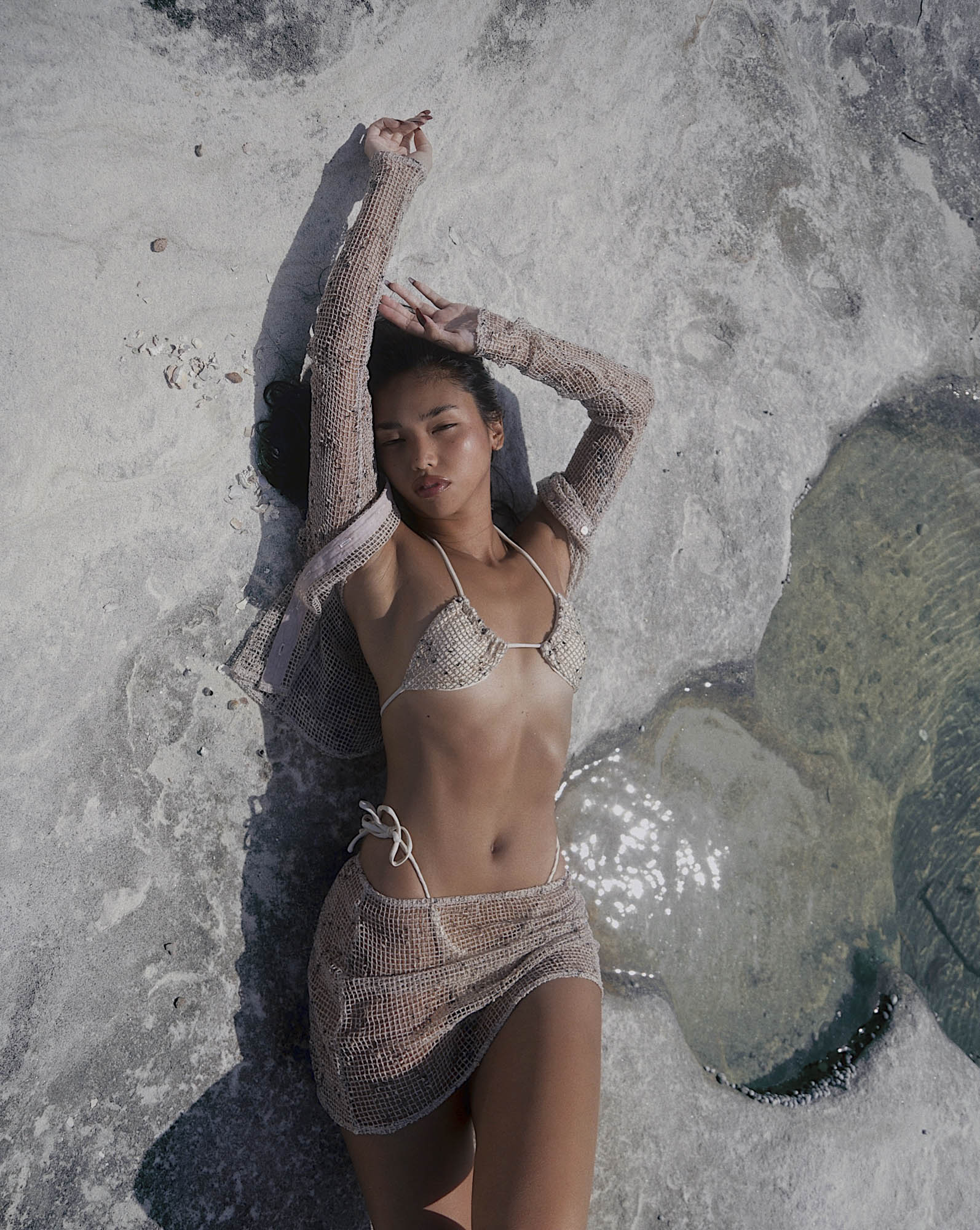 Woman lies on weathered rock formation in a beige mesh bikini and matching coverup with long sleeves, posing with arms raised.