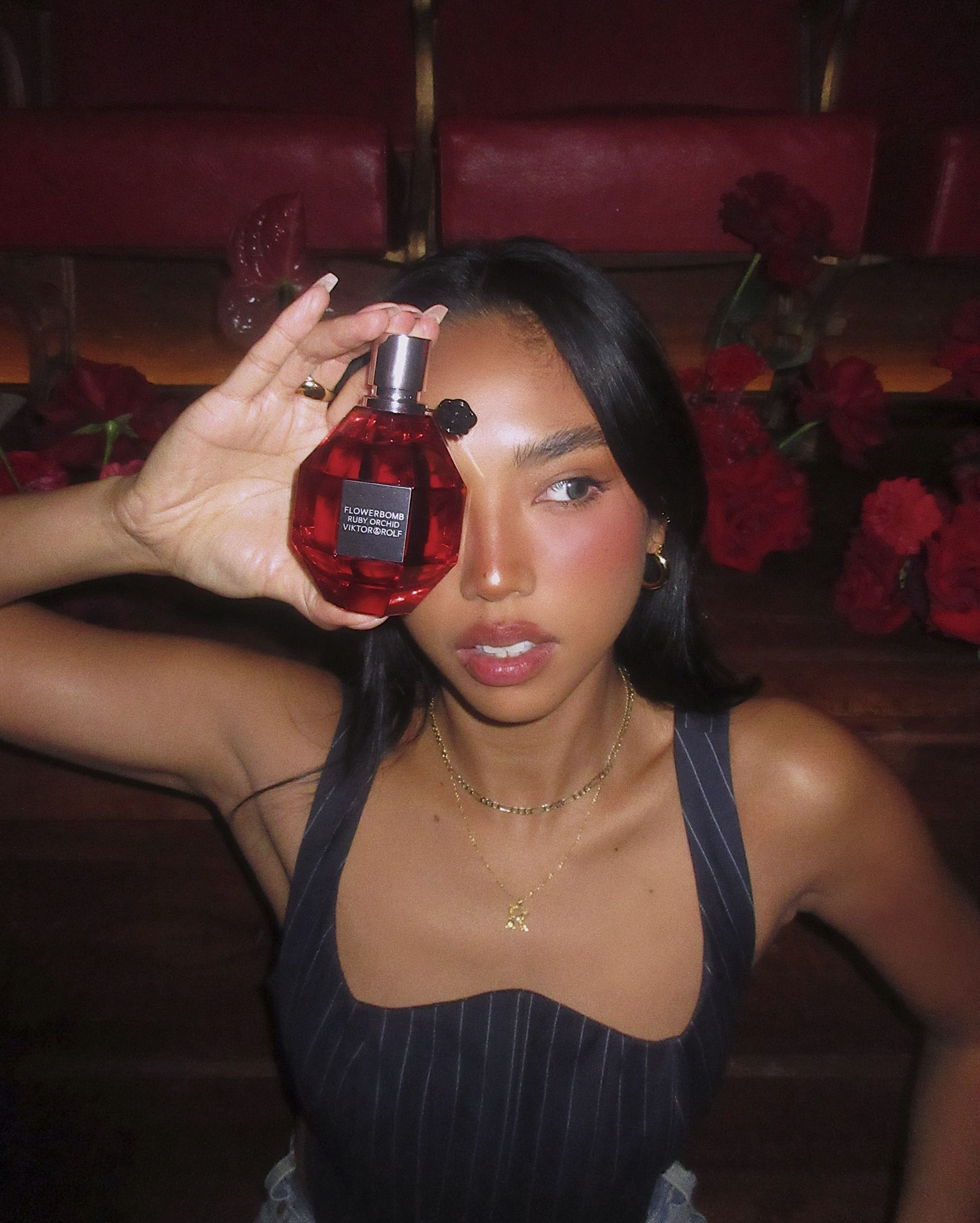Young woman poses indoors with a red perfume bottle in hand near red roses, wearing a pinstripe top and delicate gold jewelry.