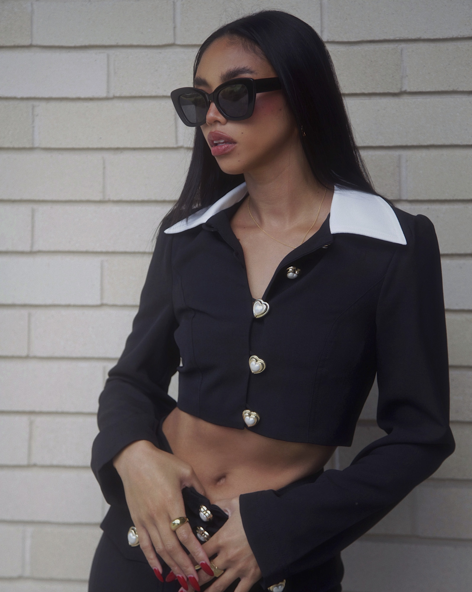 A young woman poses in a black crop top suit and sunglasses with brick background. Heart-shaped button details accent her chic look.