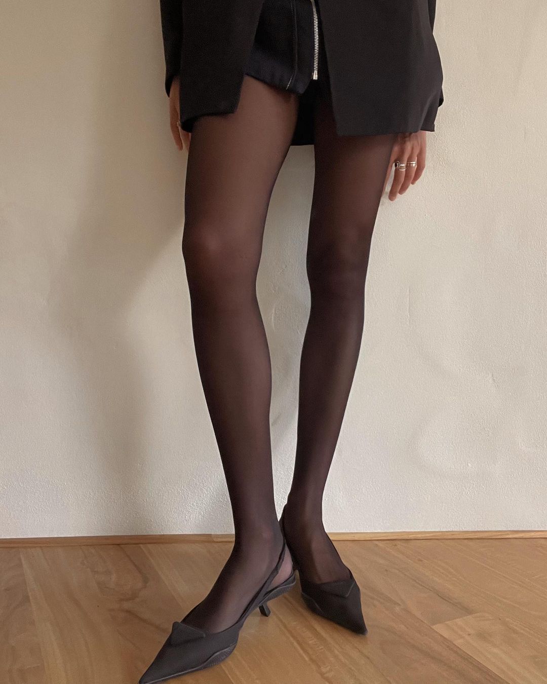 A standing woman showcases a black blazer, shorts, sheer black tights, and pointed-toe kitten heels on a wood floor against a cream-colored wall.