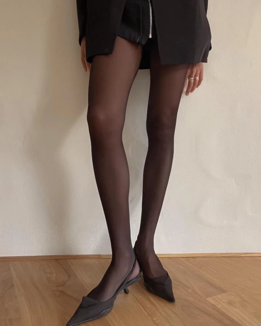A standing woman showcases a black blazer, shorts, sheer black tights, and pointed-toe kitten heels on a wood floor against a cream-colored wall.