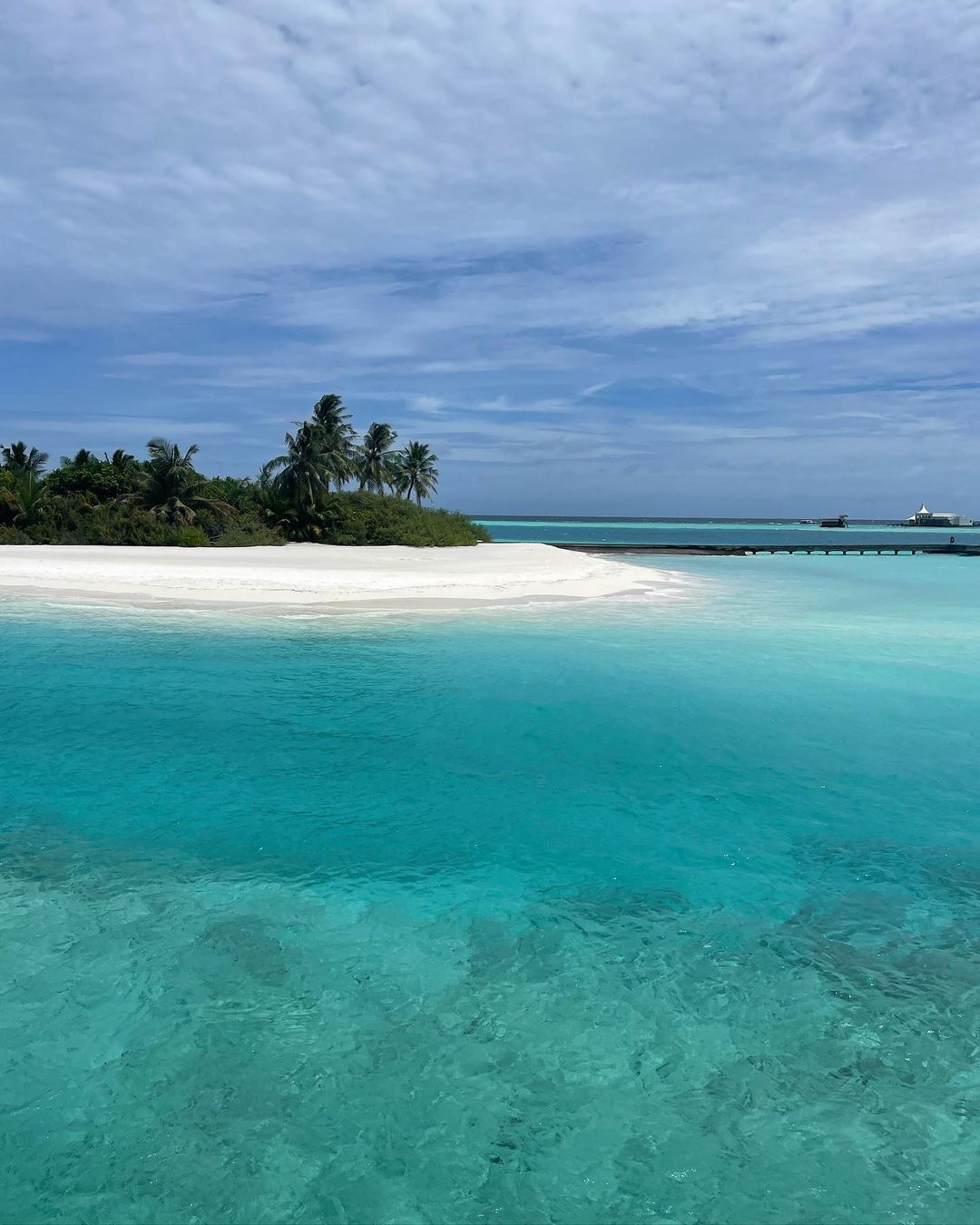Tropical island paradise featuring a white sandy beach, turquoise water, palm trees, and a blue sky with scattered clouds on a sunny day.