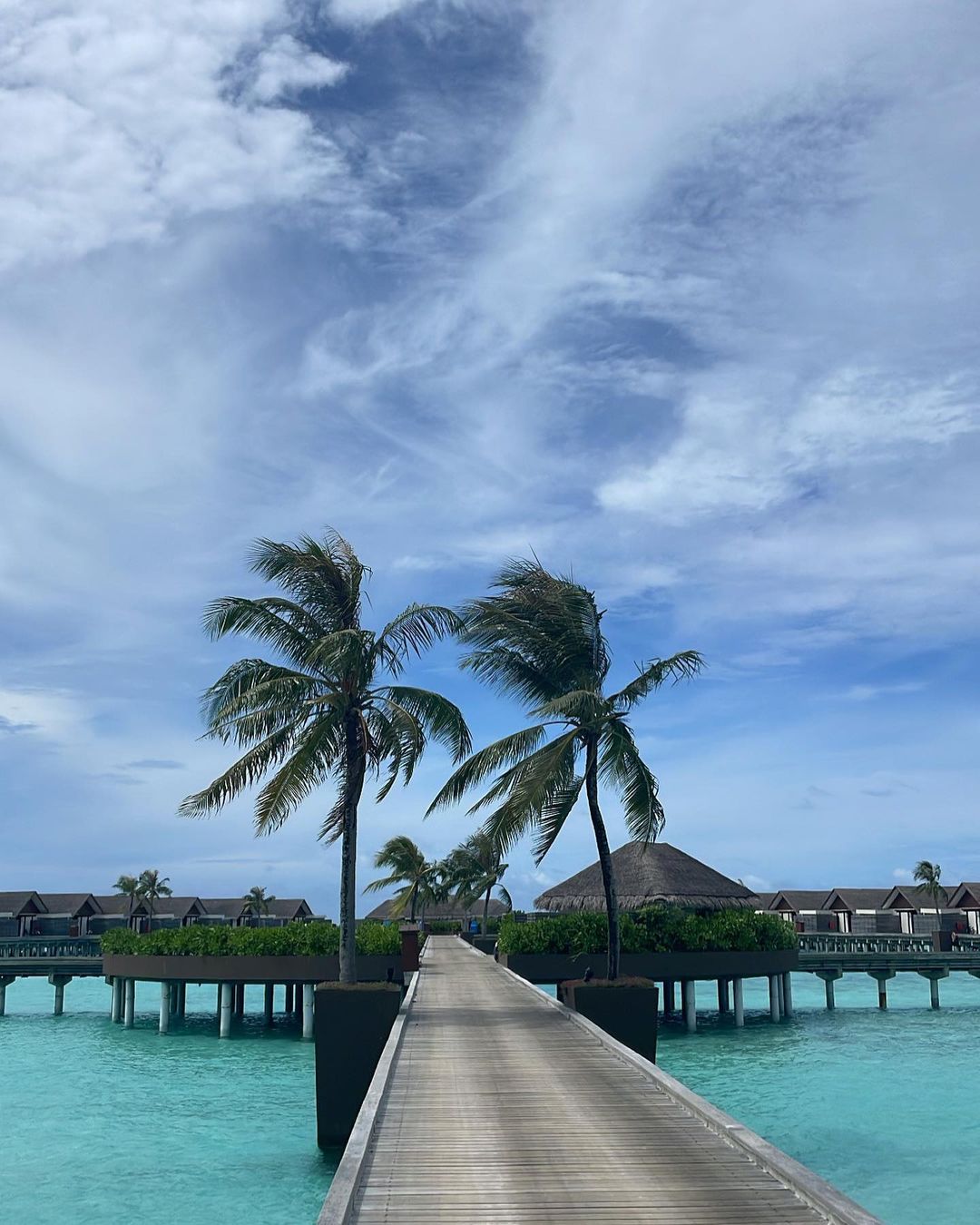 Scenic view of a wooden boardwalk leading to tropical stilt houses over turquoise water under a bright blue sky with clouds.