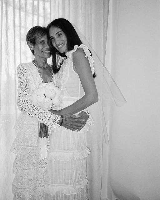 Black and white image of two women, one elderly and one young, embracing indoors in light-colored dresses; the younger holds a bouquet.