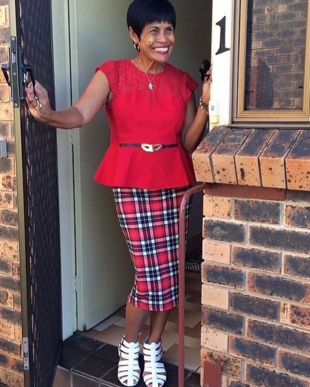 Smiling elderly African-descent woman in a red top and plaid skirt stands in a doorway with a walking stick outside a brick building.