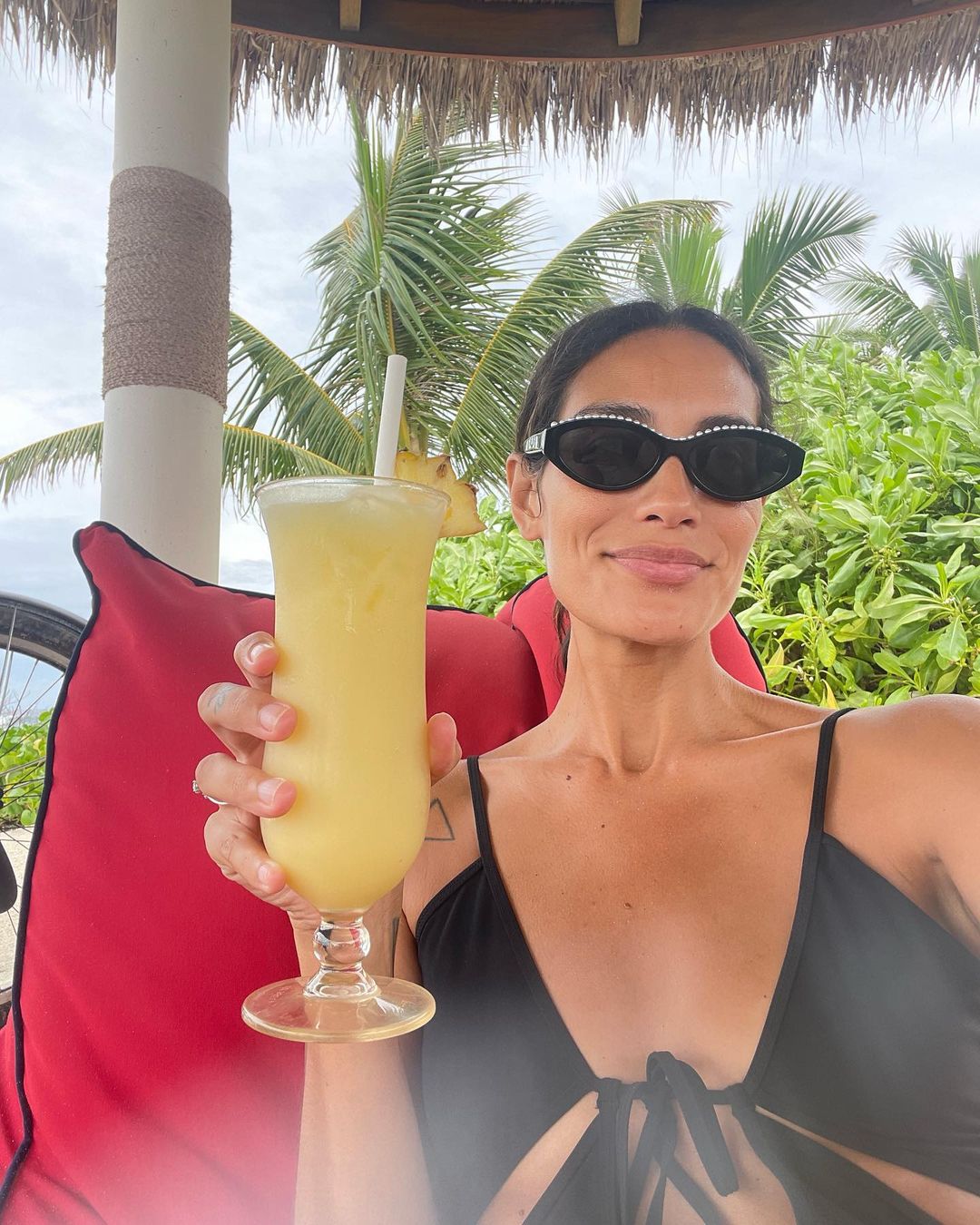 Adult female in sunglasses holds tall yellow drink at tropical beach resort with palms, red cushion. Sunny, relaxed vacation vibes.