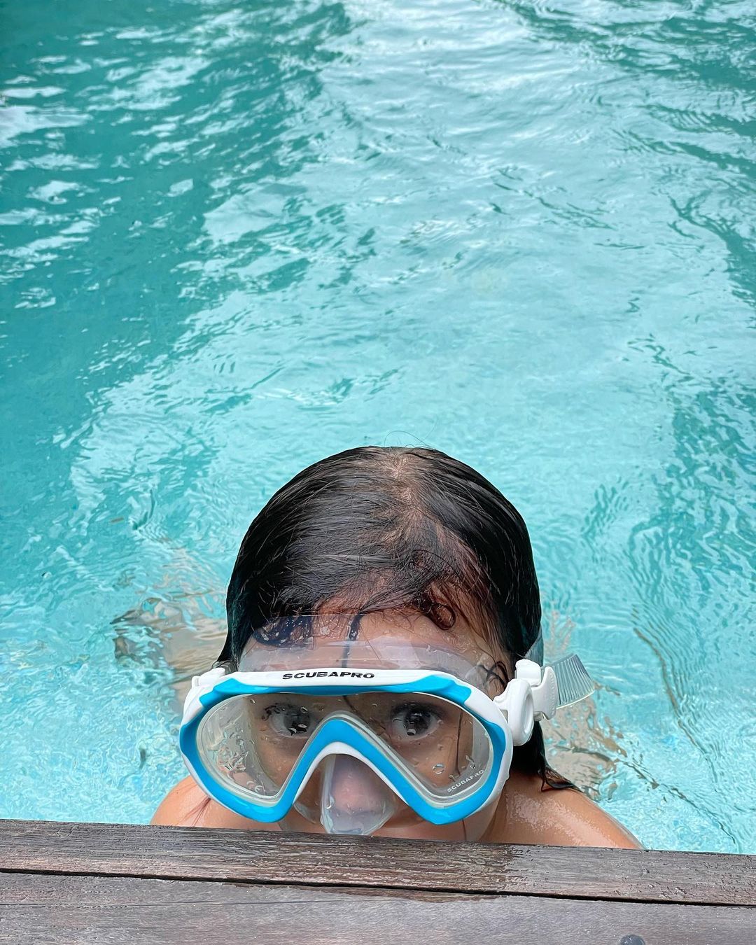 A person swimming in a pool with a blue and white SCUBAPRO diving mask, looking at the camera with a wooden poolside edge visible.