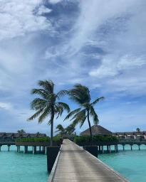A wooden boardwalk extends over bright turquoise water between two tropical resort villas, flanked by palm trees under a cloudy blue sky.