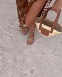 Close-up shot of a person standing on a sandy beach wearing a patterned brown skirt, strappy tan sandals, and holding a straw basket bag.