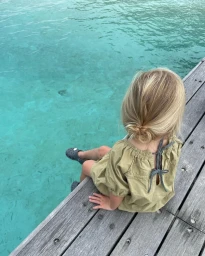 A young child with blonde hair wearing an olive dress sitting on a wooden dock above clear turquoise water.