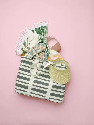 Overhead studio shot of a striped beach bag with a tropical beach towel, visor, water bottle, and flip-flops on a pink background.