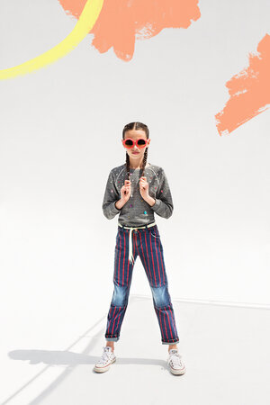 Full shot of a young girl standing in a studio wearing fashionable jeans, a gray sweatshirt, pink sunglasses, and white sneakers.