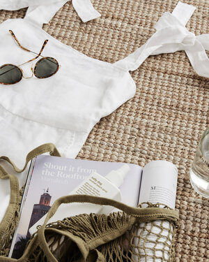 Summer flat lay of a white linen top, sunglasses, a bag, and summer essentials on a woven rug.  