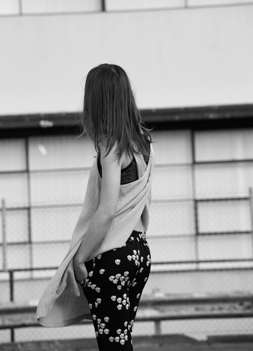 Back view of a young woman with long hair wearing patterned leggings and a long top, standing in an indoor location with daylight.