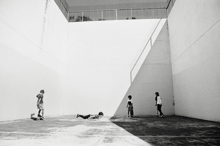 Black and white photograph of four children playing in a concrete enclosure, one on a skateboard, one on the ground, and two with scooters.