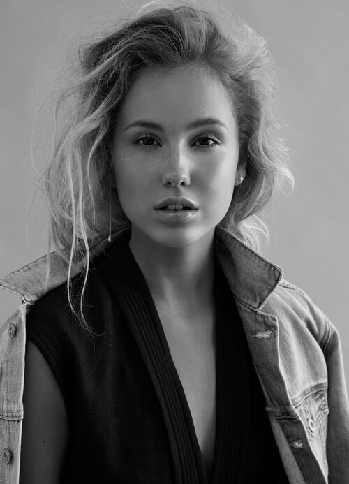 Black and white portrait of an adult woman with blonde curly hair, wearing a denim jacket over a low-cut dress, indoors.