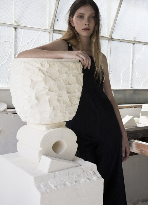 Young woman in a black dress leans against a textured white art sculpture inside a sunlit studio with large windows, a minimalist aesthetic.
