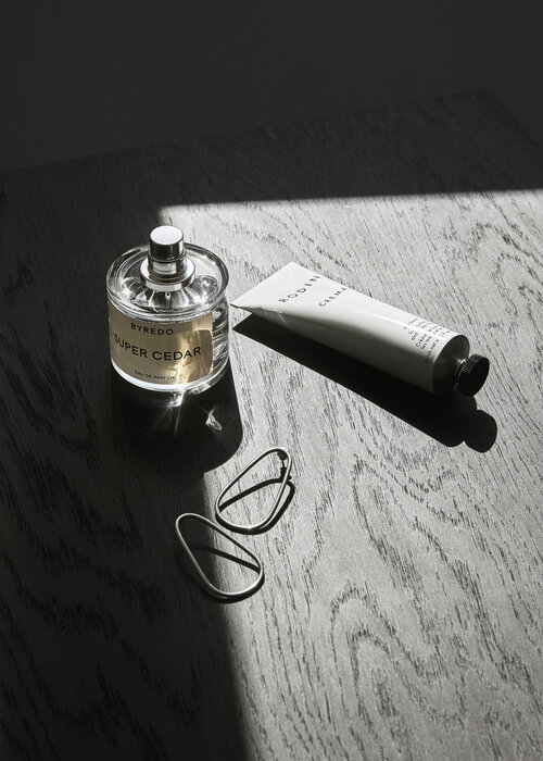 Still life of Byredo perfume bottle, lotion tube, and metal earrings on a dark wooden surface with natural light and shadows.